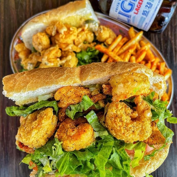 A fried shrimp po'boy sandwich with lettuce, fries, and a beer bottle on a metal tray.